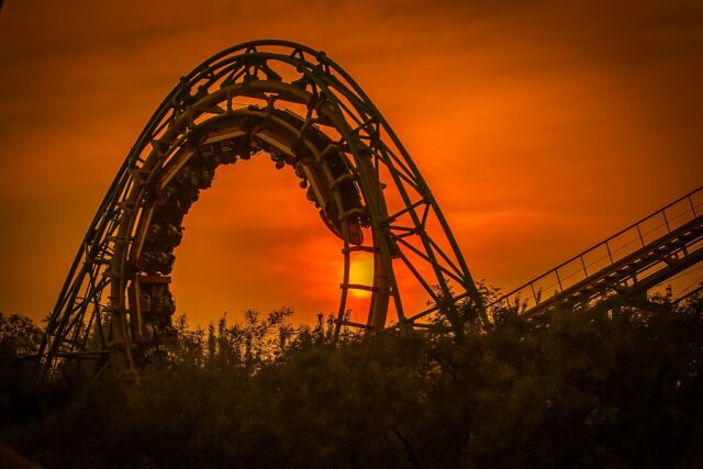 A roller coaster train goes upside down through a loop against an orange California sunset sky, with trees silhouetted in the foreground—a thrilling reminder of what to do at an amusement park.