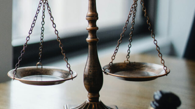 A brass balance scale sits on a wooden table beside a gavel, symbolizing how legal decisions—and the risk of a criminal record—can have long-term consequences, with a window in the background.