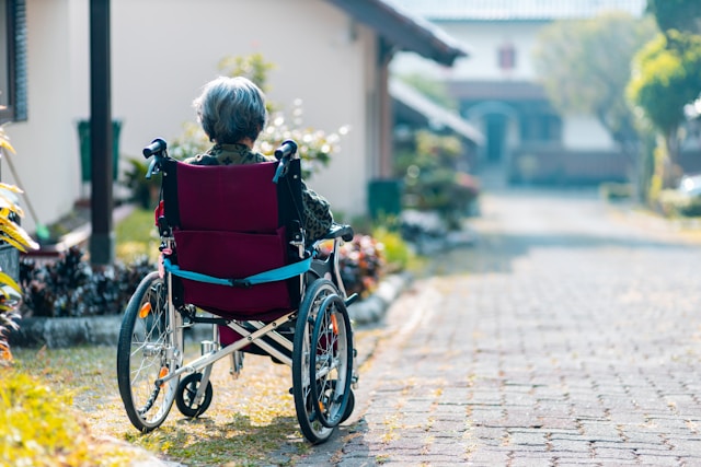 An elderly person with gray hair sits in a wheelchair on a paved path outside in California, facing away from the camera toward houses and greenery.