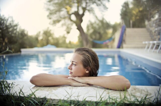 Person with wet hair resting arms on the edge of an outdoor swimming pool, looking to the side; trees and a hammock are visible in the background—a reminder to keep pool safety in mind, especially during Labor Day weekend.